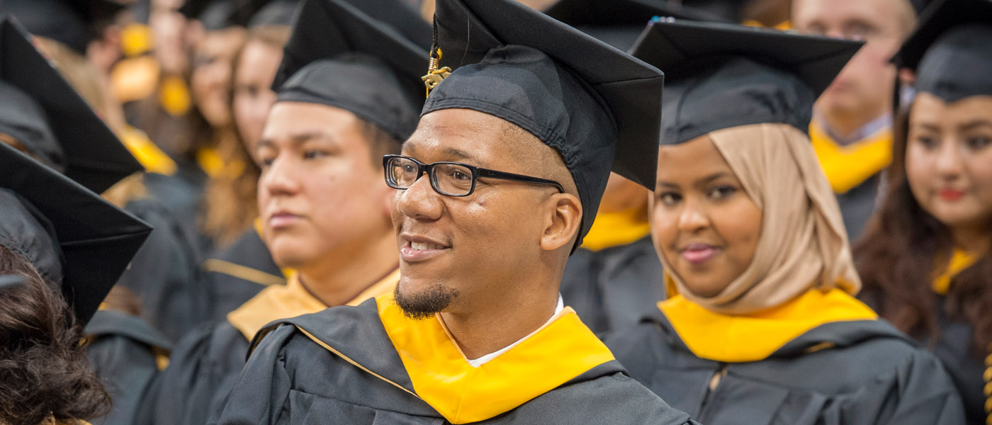 A crowd of graduates in caps and gowns. A male graduate in glasses smiles in the foreground.