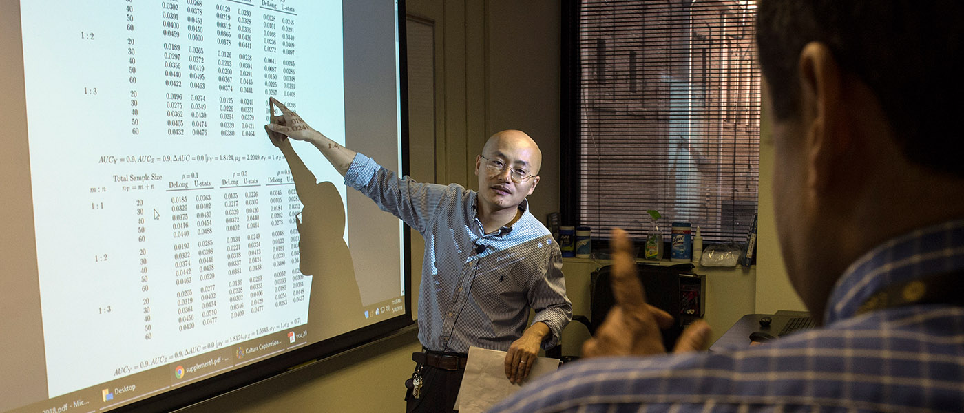 A male teacher stands in front of a white board with a project image of data in a table
